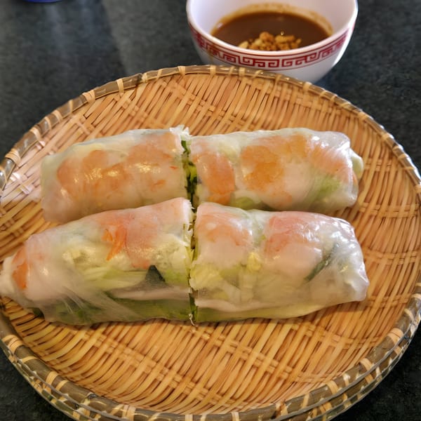 Four Vietnamese spring rolls with shrimp and vegetables on a woven plate, with dipping sauce.