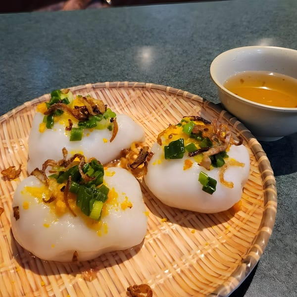 Three Vietnamese Banh Beo dumplings with green onions and crispy shallots on a woven plate with sauce.