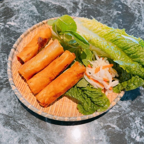 Four golden-brown Vietnamese spring rolls with fresh lettuce, mint, and pickled vegetables on a woven plate.